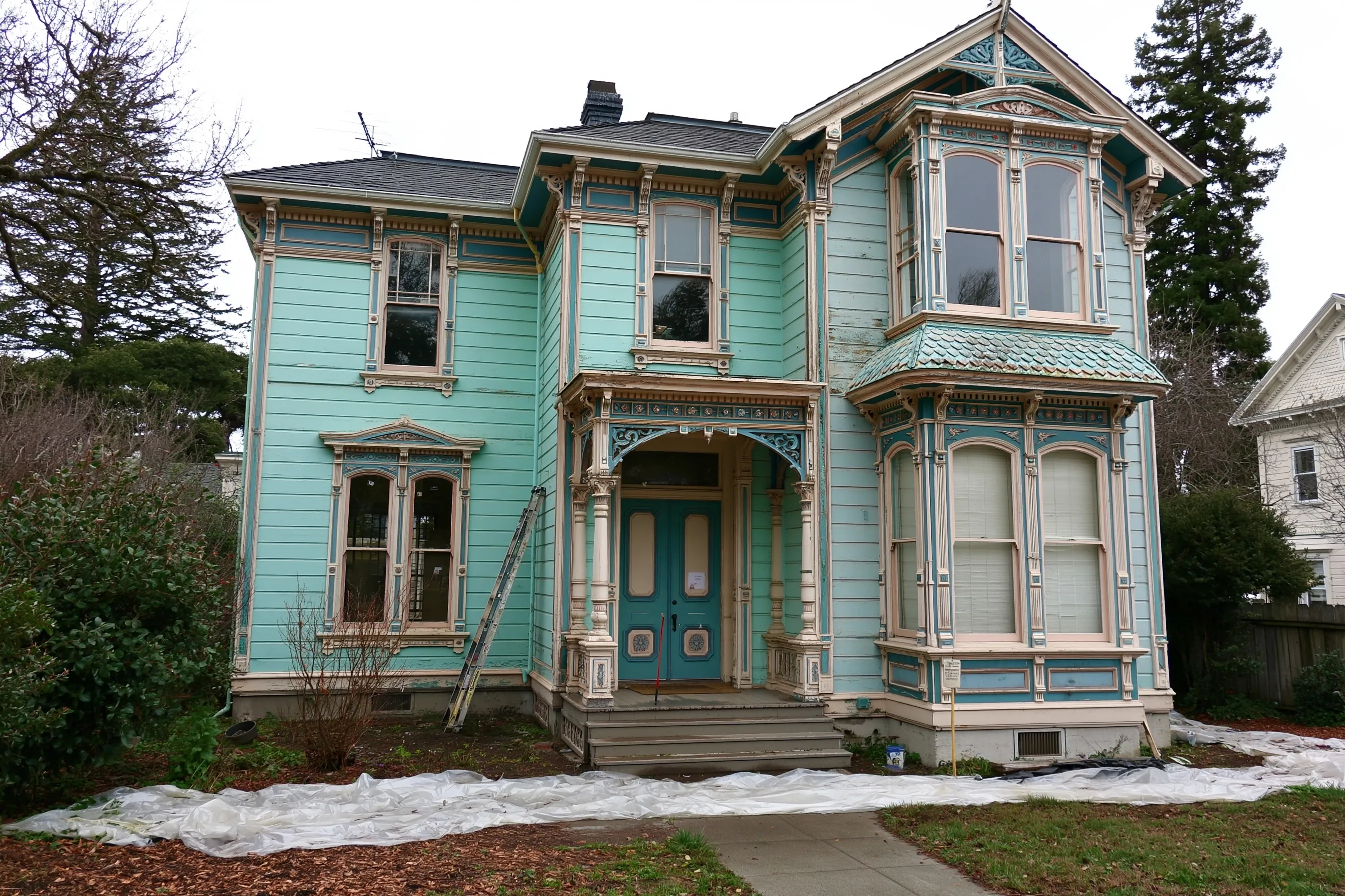 Two-story Victorian house with painted wood siding and ornate trim, a ladder against the porch and plastic sheeting on the ground in front